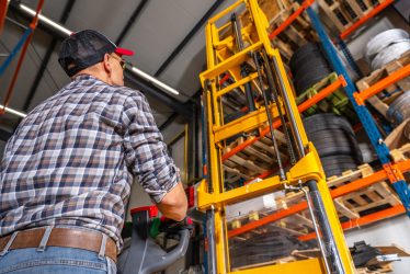 A warehouse worker operates a forklift to retrieve tires stacked on shelves in a storage area. The worker is focused and wearing safety gear, surrounded by organized shelving filled with various equipment and materials.